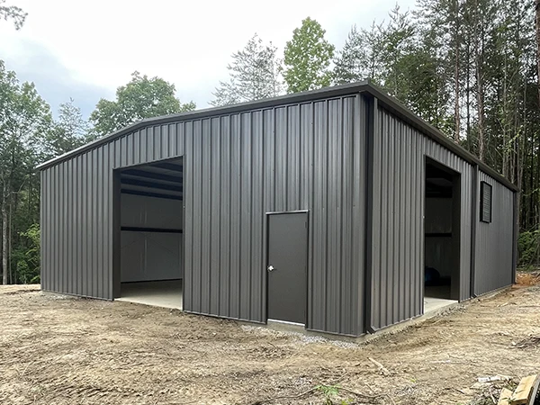 A gray metal storage building stands on a cleared lot, surrounded by trees, with a side door and open bays.