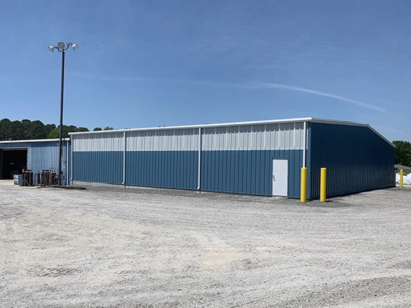 A large blue metal building with a light pole and yellow posts, set against a clear sky and gravel lot.