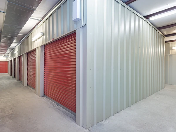 Interior of a storage facility showing several red roll-up doors, beige metal walls, and overhead lighting along a spacious aisle.