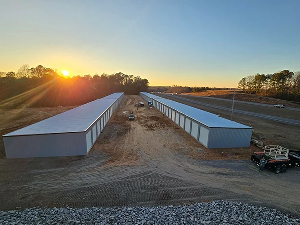 Two rows of storage units line a dirt lot near a highway, illuminated by a vibrant sunset in the background.